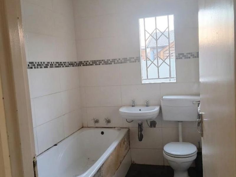 A functional bathroom featuring a bathtub, wall-mounted basin, and toilet, with tiled finishes and a security-gated window.