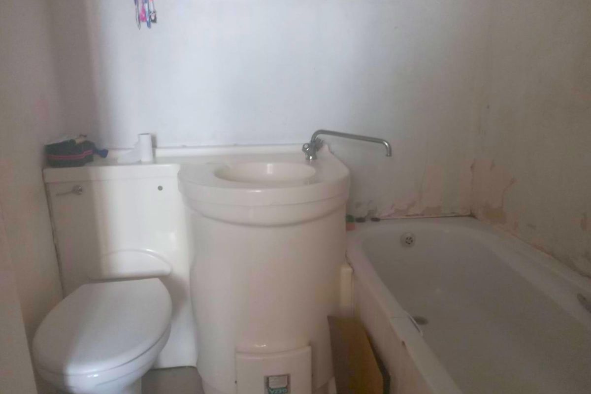 Compact bathroom featuring an integrated toilet and basin unit alongside a standard bathtub, requiring cosmetic updates and wall repairs.