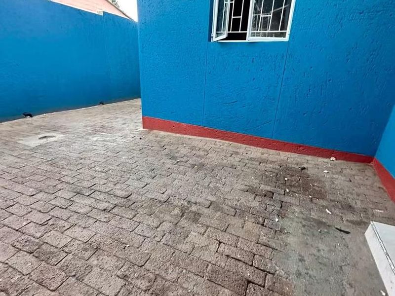 A rear exterior view of the property featuring a paved courtyard area enclosed by a blue boundary wall with security bars on the window.