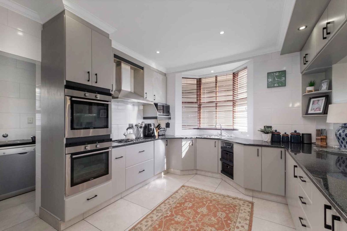 Modern kitchen featuring sleek grey cabinetry, granite countertops, built-in double ovens, stainless steel extractor fan, and a bright bay window with fitted blinds.