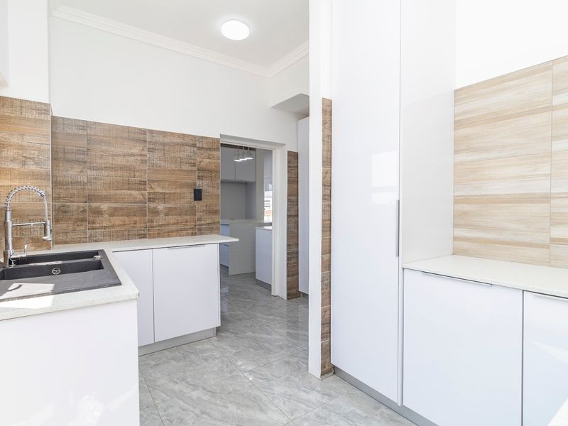 Modern kitchen featuring white cabinetry, stone countertops, wood-look tiled backsplash, and high-quality floor tiles, with a clean, minimalist aesthetic.