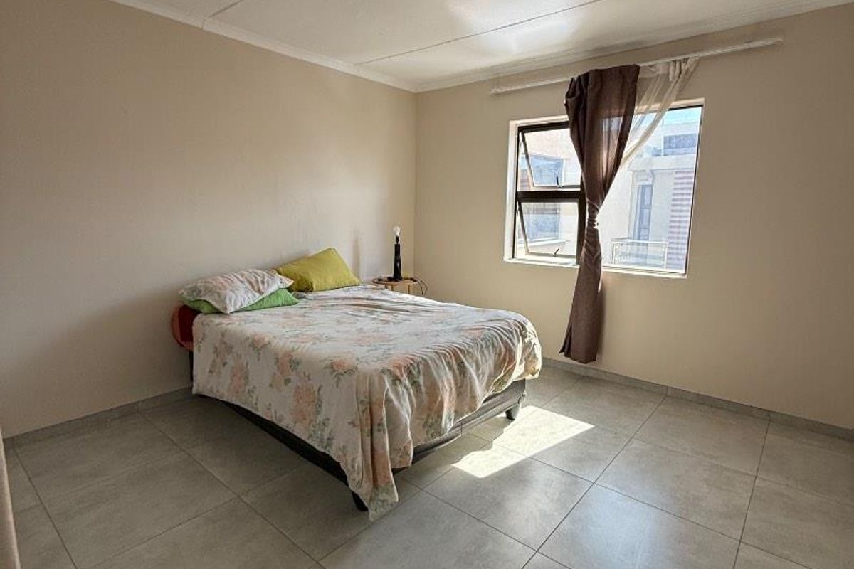 A simple bedroom featuring neutral wall tones, large format floor tiles, and an aluminium window frame providing natural light.