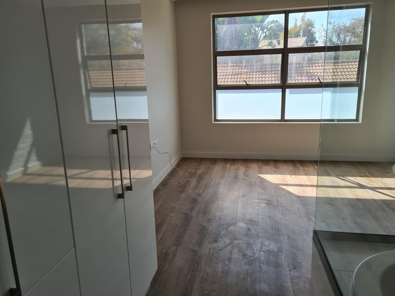 A bright, empty bedroom featuring modern laminate flooring, built-in cupboards, and large aluminium-framed windows allowing for ample natural light.