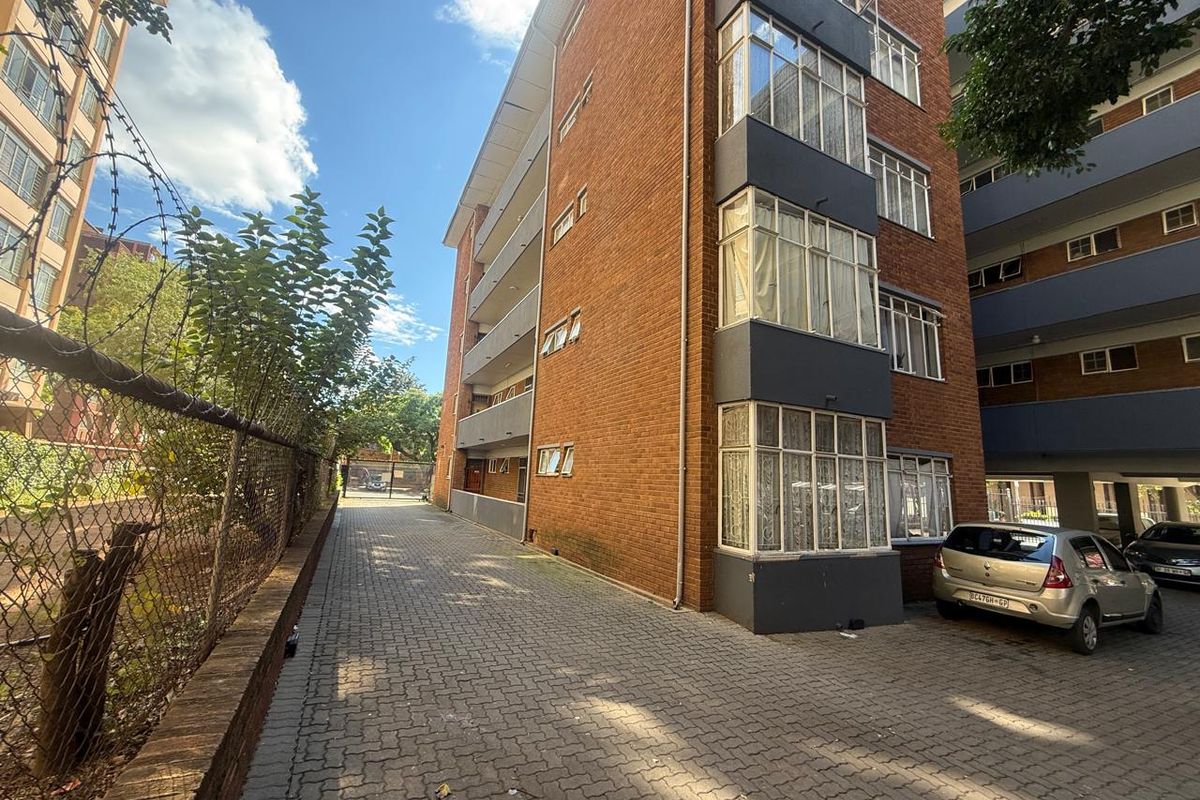 Exterior view of a multi-story brick apartment building featuring paved driveway access and security fencing.