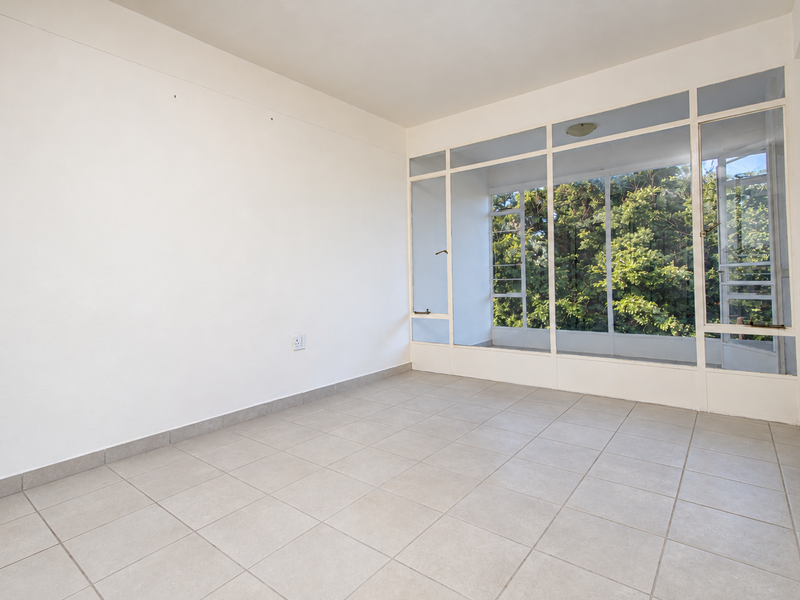 A bright, empty living area featuring neutral tiled flooring and a large glass window wall providing natural light and garden views.