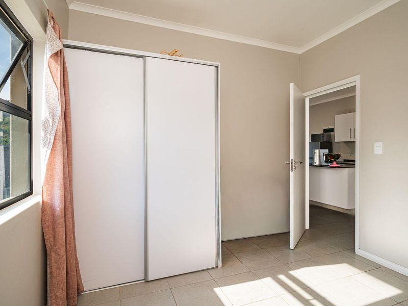 A bright, empty bedroom featuring tiled flooring, a built-in sliding door wardrobe, and a top-hung window, with a doorway leading to the kitchen area.