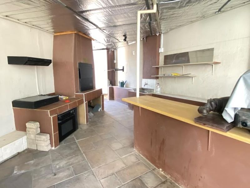 Covered outdoor entertainment area featuring a built-in braai, oven, bar counter, and sink, with stone tile flooring and exposed insulation ceiling.