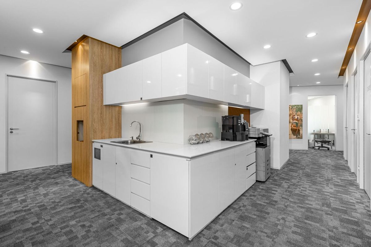 Modern office kitchenette featuring sleek white cabinetry, integrated sink, and wood-panelled accent column, set within a professional workspace with recessed lighting.
