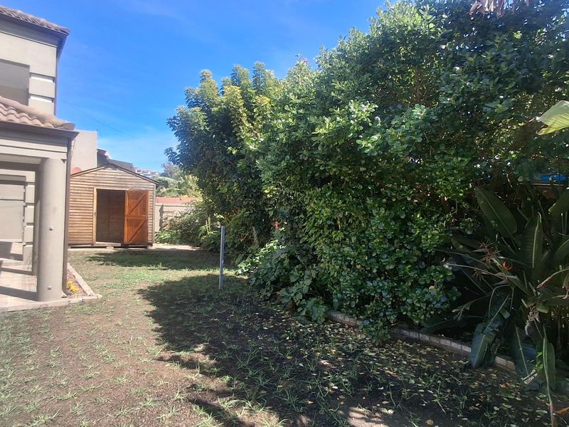 A rear garden area featuring a tiled patio, a wooden storage shed, and established garden beds with mature shrubs.