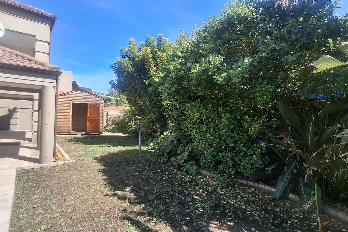 A rear garden area featuring a tiled patio, a wooden storage shed, and established garden beds with mature shrubs.
