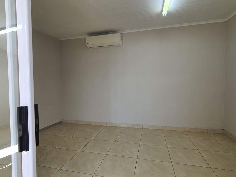 A clean, empty office space featuring tiled flooring, a wall-mounted air conditioning unit, and a sliding glass entrance door.