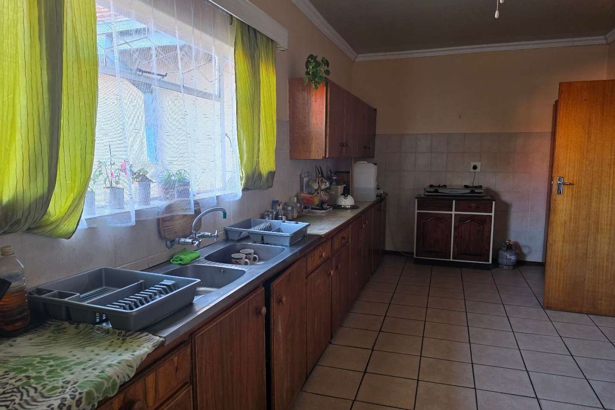 A functional kitchen featuring wooden cabinetry, a double basin stainless steel sink, tiled splashbacks, and tiled flooring, offering potential for modernization.