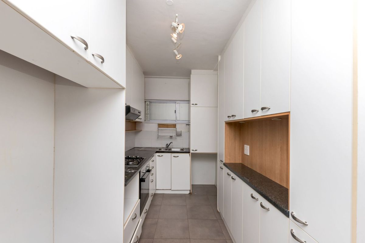 A galley-style kitchen featuring white cabinetry, dark stone countertops, a built-in gas hob, oven, and stainless steel sink.