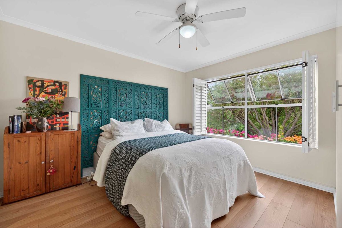 A bright bedroom featuring laminate flooring, a ceiling fan, and awning windows with plantation shutters overlooking a garden.