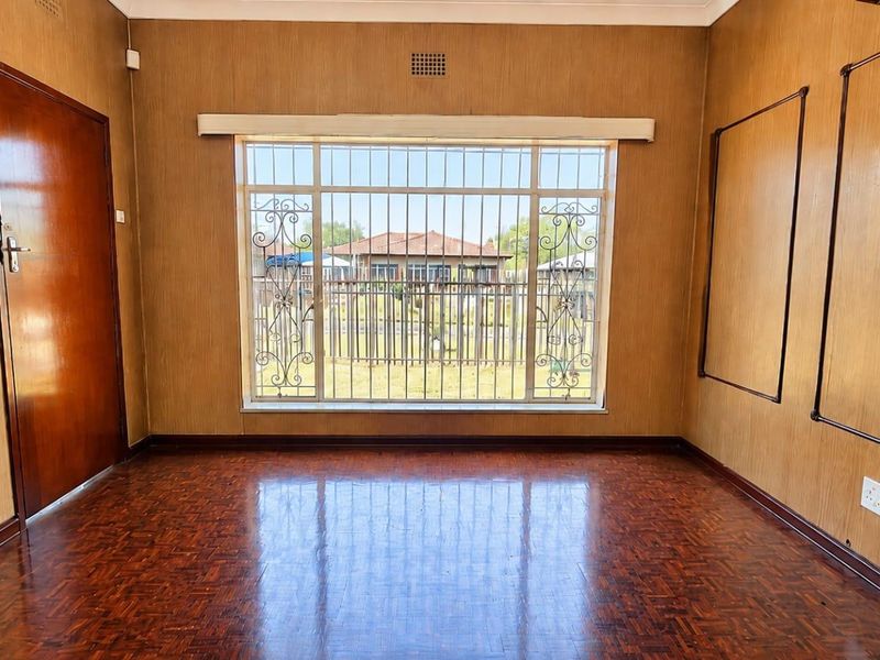 An empty living room featuring classic parquet flooring, wood-paneled walls, and a large window fitted with security bars.