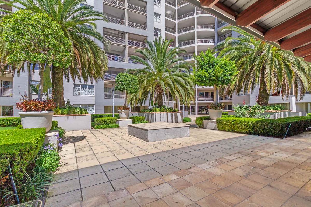 A well-maintained paved courtyard area featuring landscaped garden beds, mature palm trees, and direct access to the apartment building facade.