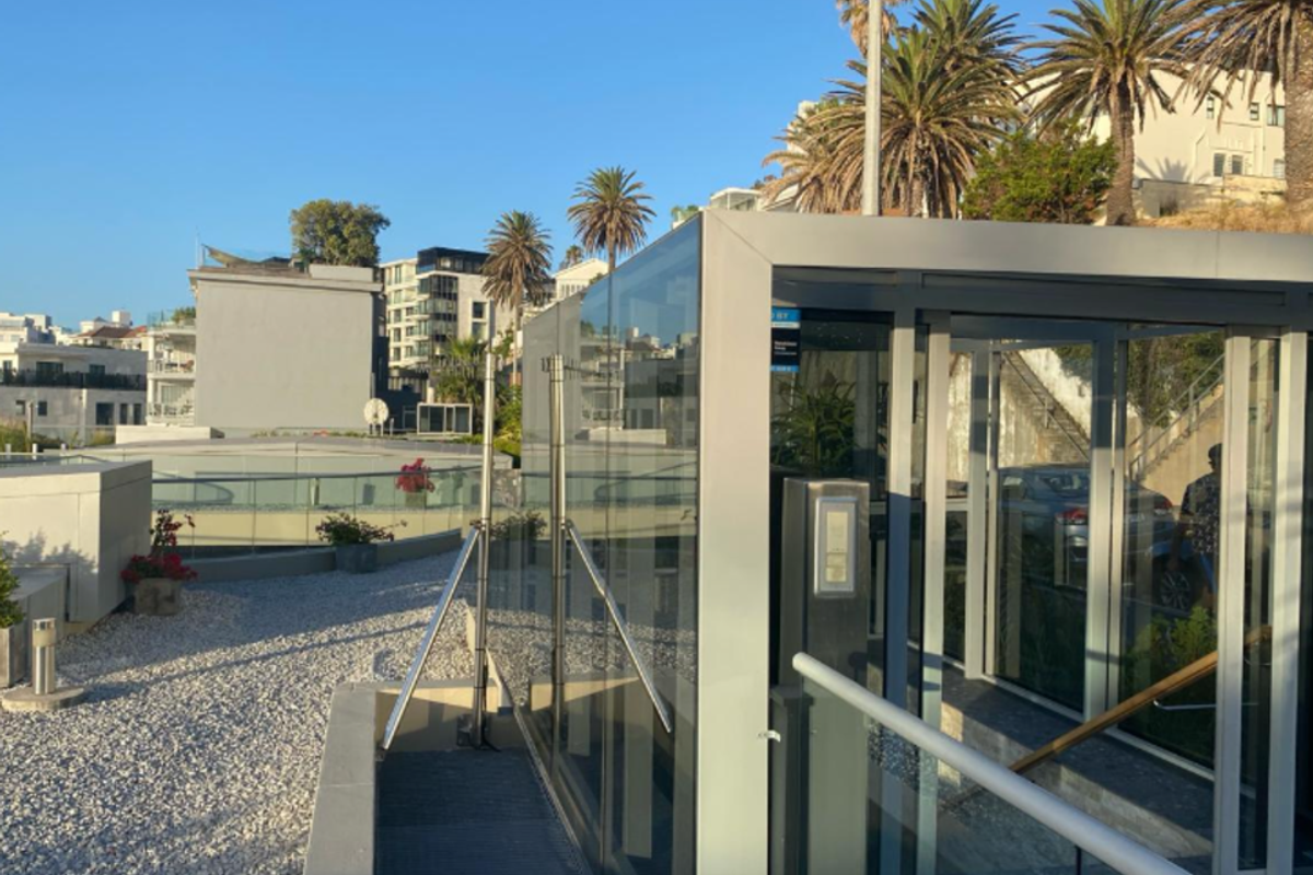 A modern outdoor terrace featuring a glass elevator shaft, gravel landscaping, and a glass balustrade overlooking the surrounding apartment complex.