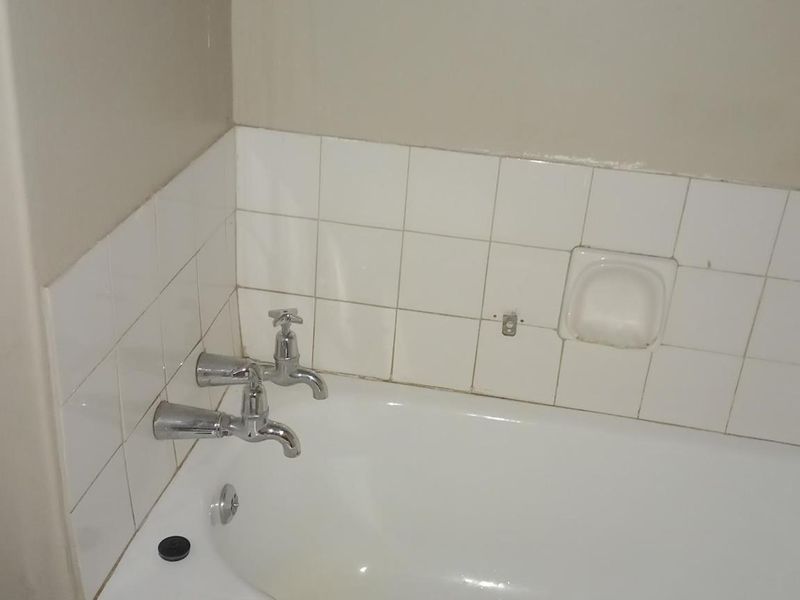 A basic bathroom featuring a standard bathtub with chrome faucets and white wall tiling, currently showing signs of wear and missing fixtures.