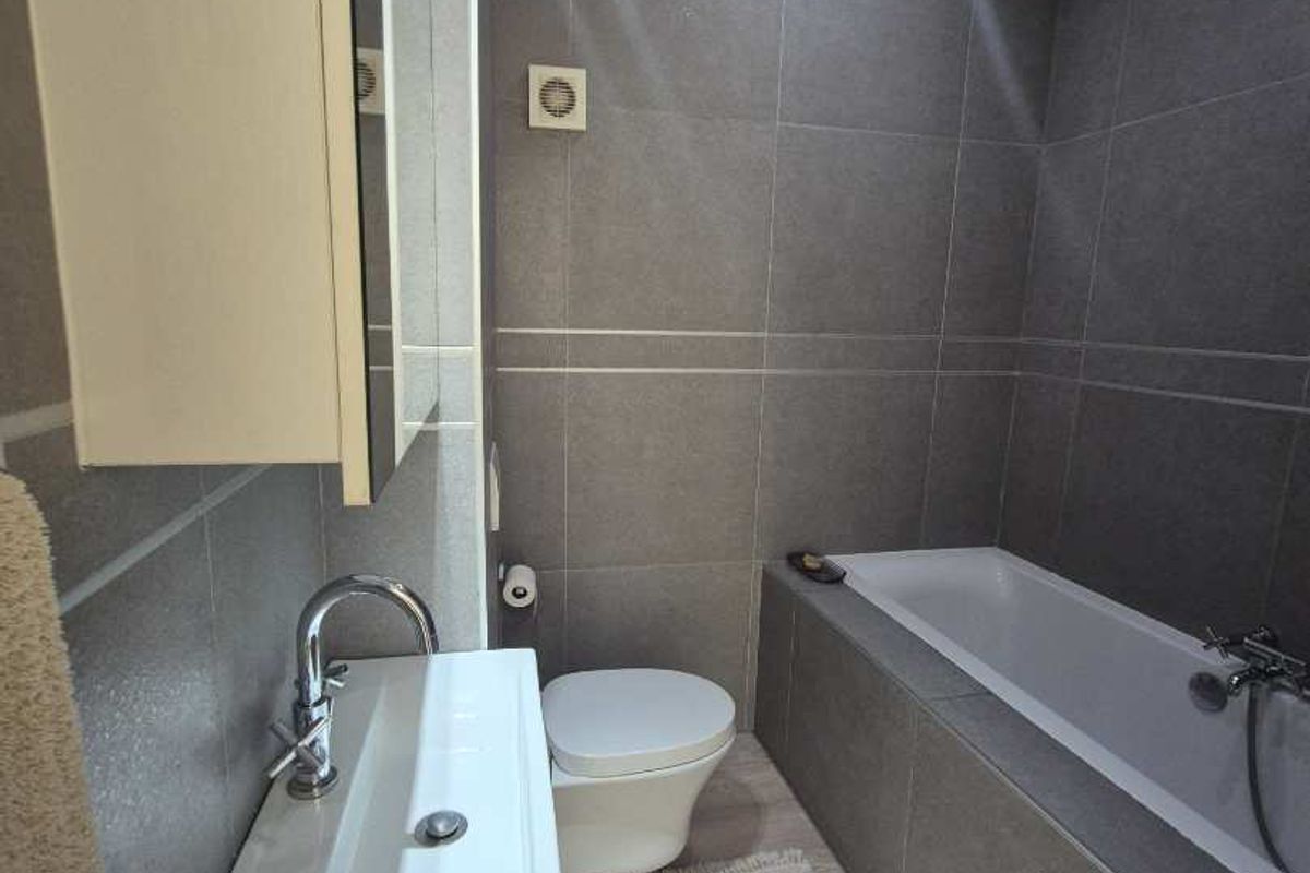 Modern bathroom featuring grey wall tiles, a built-in bathtub, a wall-mounted toilet, and a sleek vanity basin with chrome fixtures.