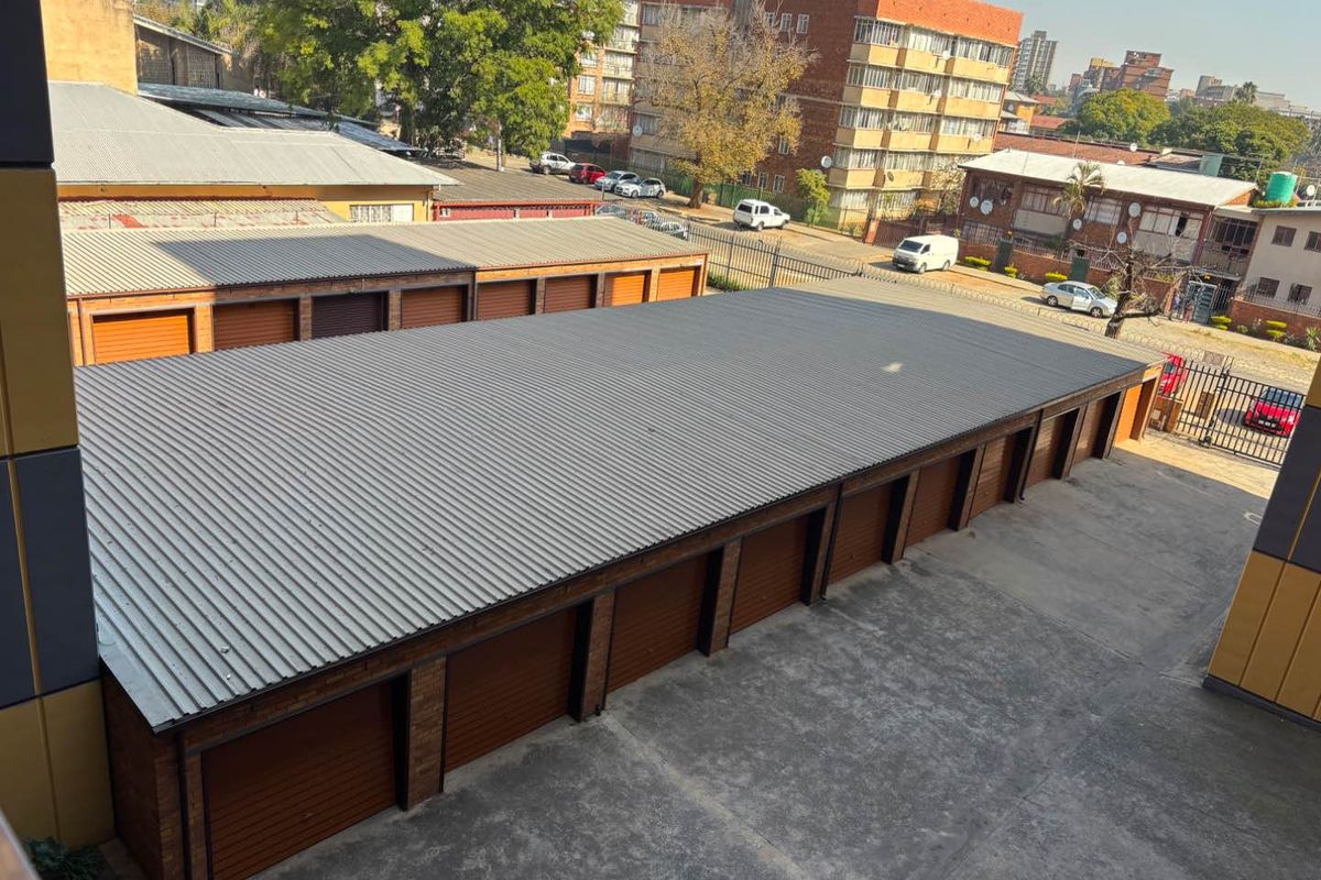 An elevated view of a row of brick garages featuring corrugated metal roofing and multiple roller shutter doors with a concrete driveway.