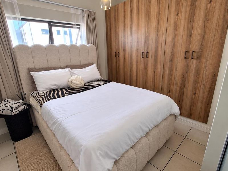 A well-maintained bedroom featuring modern wood-finish built-in cupboards, neutral tiled flooring, and a contemporary pendant light fixture.