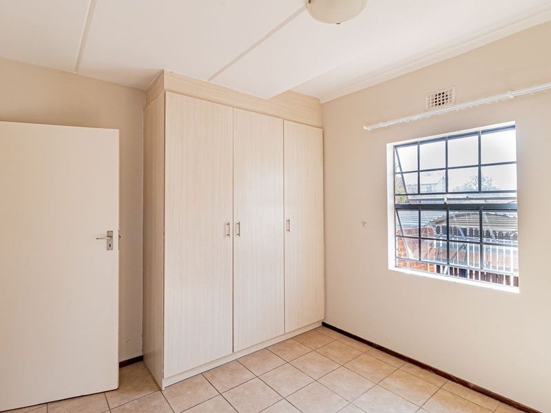 A clean, empty bedroom featuring neutral tiled flooring, built-in light wood-finish cupboards, and a steel-framed window providing natural light.