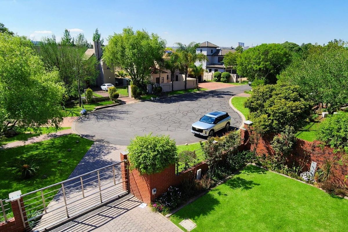 An elevated aerial view of the property entrance, featuring a landscaped garden, brick boundary wall, metal gate, and paved driveway in a quiet cul-de-sac.