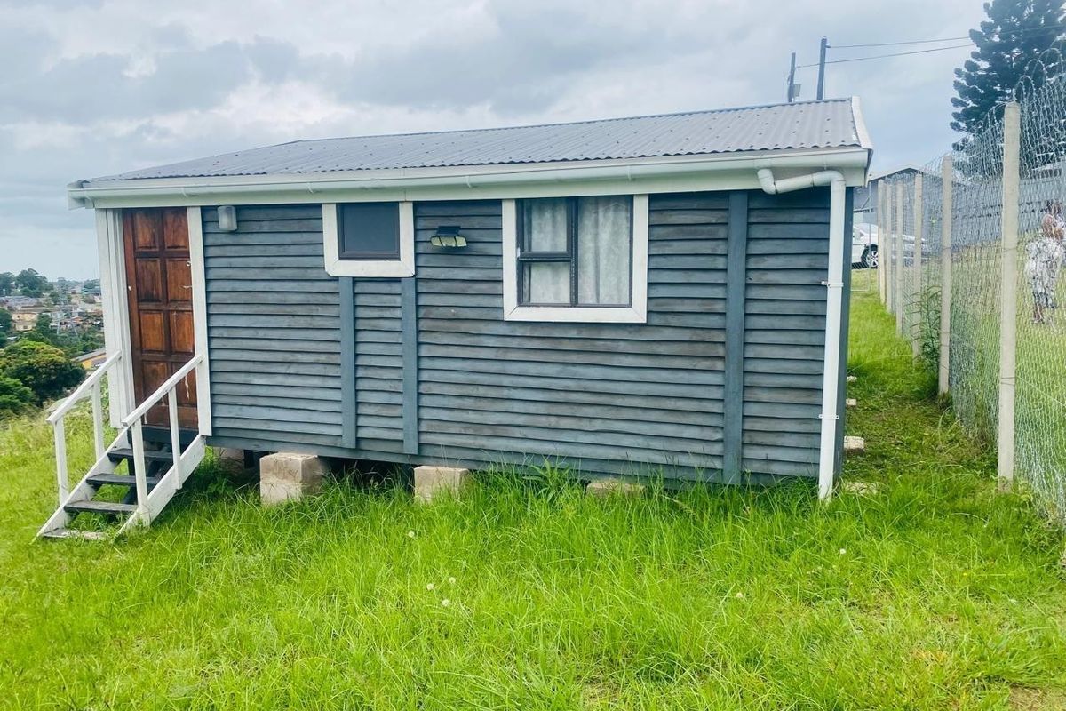 A detached timber-clad cabin structure featuring a corrugated metal roof, elevated on concrete blocks, situated on a grassy residential plot with perimeter fencing.