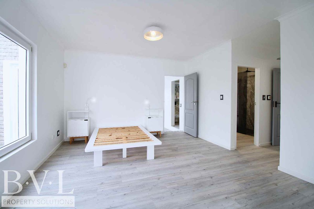 A bright, modern bedroom featuring light wood-look flooring, neutral walls, and an en-suite bathroom entrance.