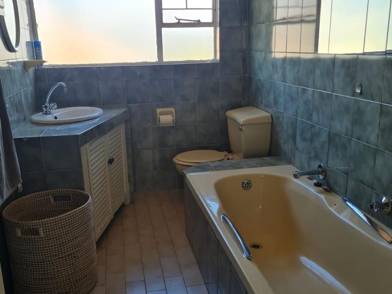 A functional bathroom featuring a built-in vanity, bathtub, and toilet with full-height wall tiling and tiled flooring.