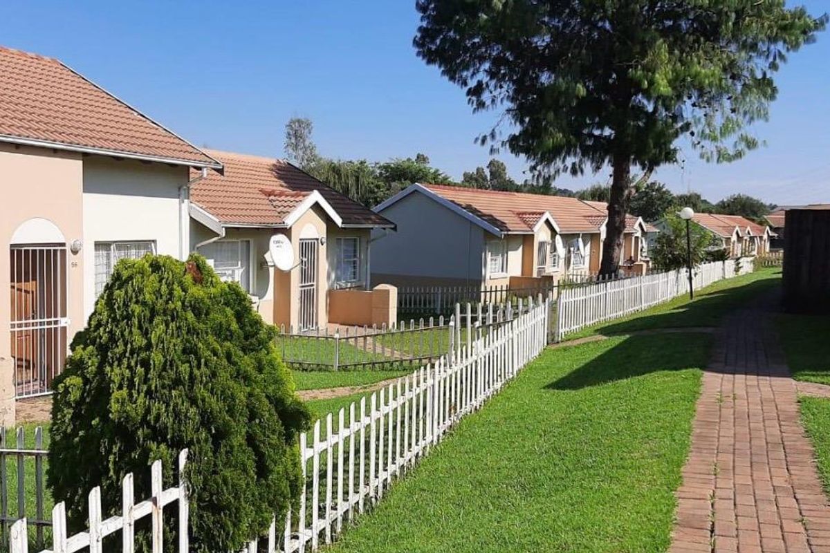 A view of a residential cluster complex featuring multiple units with tiled roofs, paved walkways, and white picket fencing in a landscaped garden setting.