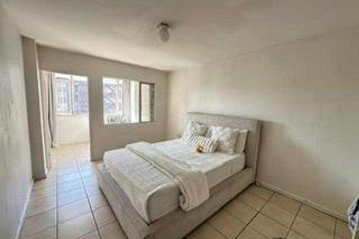 A bedroom featuring tiled flooring and a large window, with an adjacent sunroom or balcony area accessible through an open doorway.