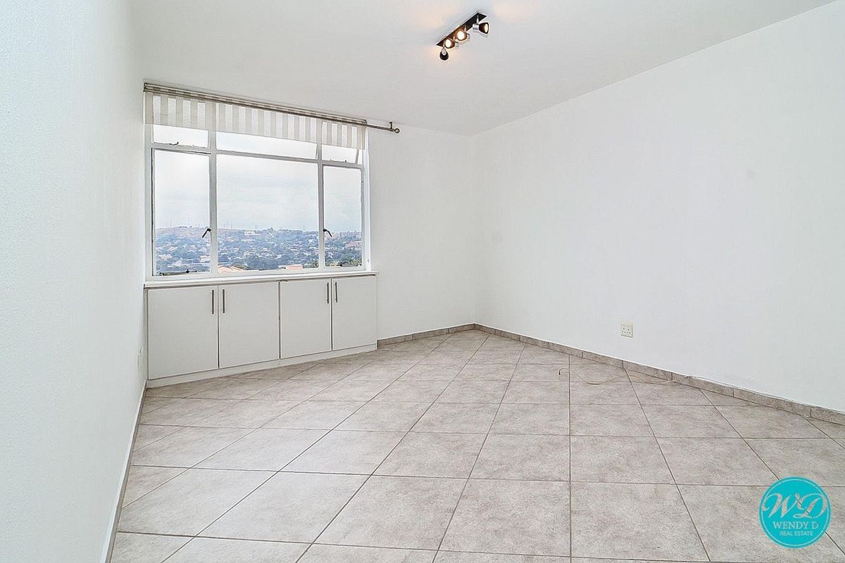 A bright, empty bedroom featuring neutral floor tiles, built-in under-window cabinetry, and a large window offering expansive elevated views.