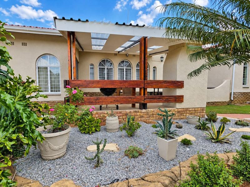 A well-maintained covered patio area featuring a timber pergola structure, brick base, and arched windows overlooking a landscaped garden.