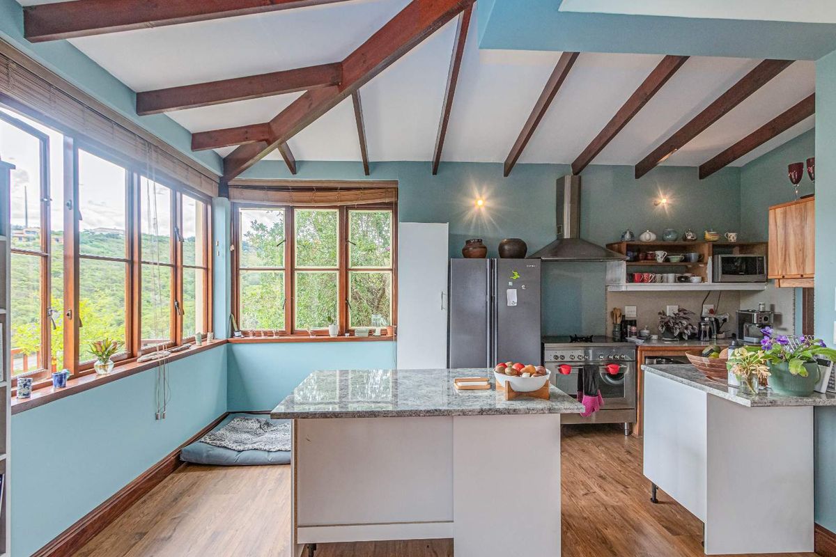 A bright kitchen featuring exposed timber ceiling beams, a central island with stone countertop, stainless steel oven, and large windows offering scenic views.