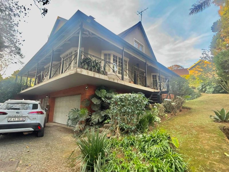 A double-story residential property featuring a wrap-around balcony, wrought iron railings, and an integrated garage, set within a landscaped garden environment.