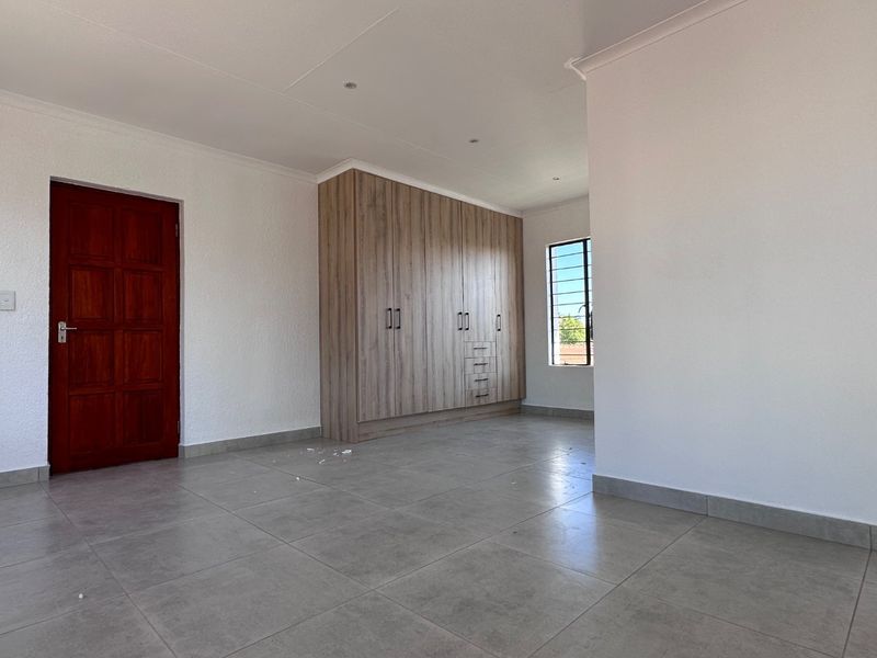 A bright, empty bedroom featuring modern tiled flooring, built-in wooden cupboards, and a window with security bars.