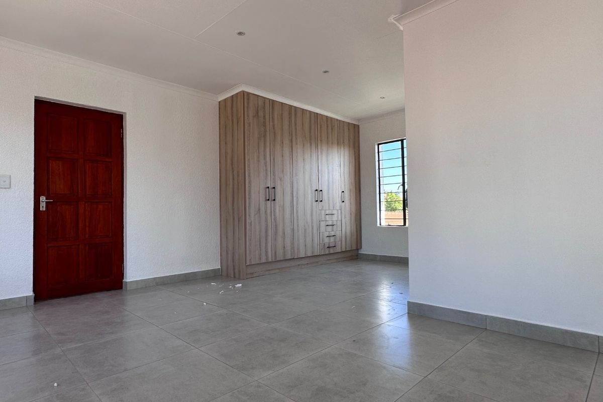 A bright, empty bedroom featuring modern tiled flooring, built-in wooden cupboards, and a window with security bars.