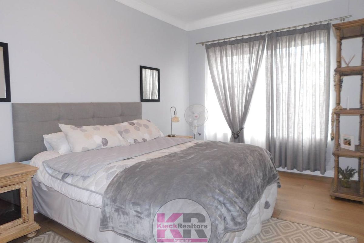 A well-maintained bedroom featuring light-toned wood flooring, neutral wall finishes, and large windows with sheer curtains allowing for natural light.