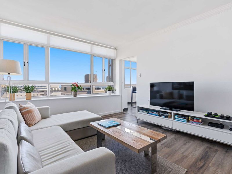 A bright and modern living room featuring large windows with city views, laminate flooring, and clean white walls.