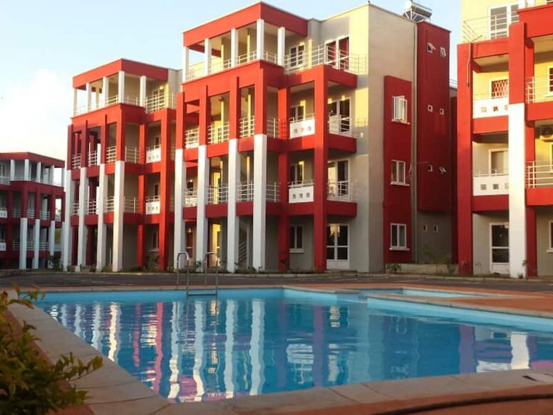 A view of a communal swimming pool set against a multi-story apartment complex featuring red and white exterior facades and private balconies.