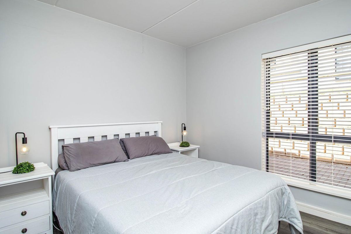 A bright and neutral bedroom featuring a large window with fitted blinds, clean wall finishes, and modern flooring.