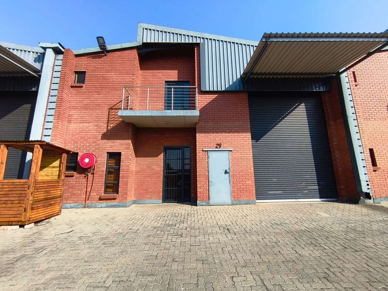 Industrial unit featuring a red brick facade, large roller shutter door, pedestrian entrance with security gate, and a small upper-level balcony.
