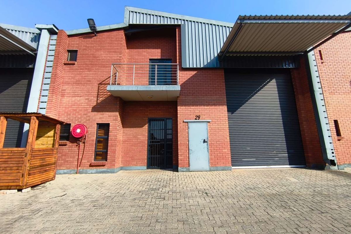 Industrial unit featuring a red brick facade, large roller shutter door, pedestrian entrance with security gate, and a small upper-level balcony.