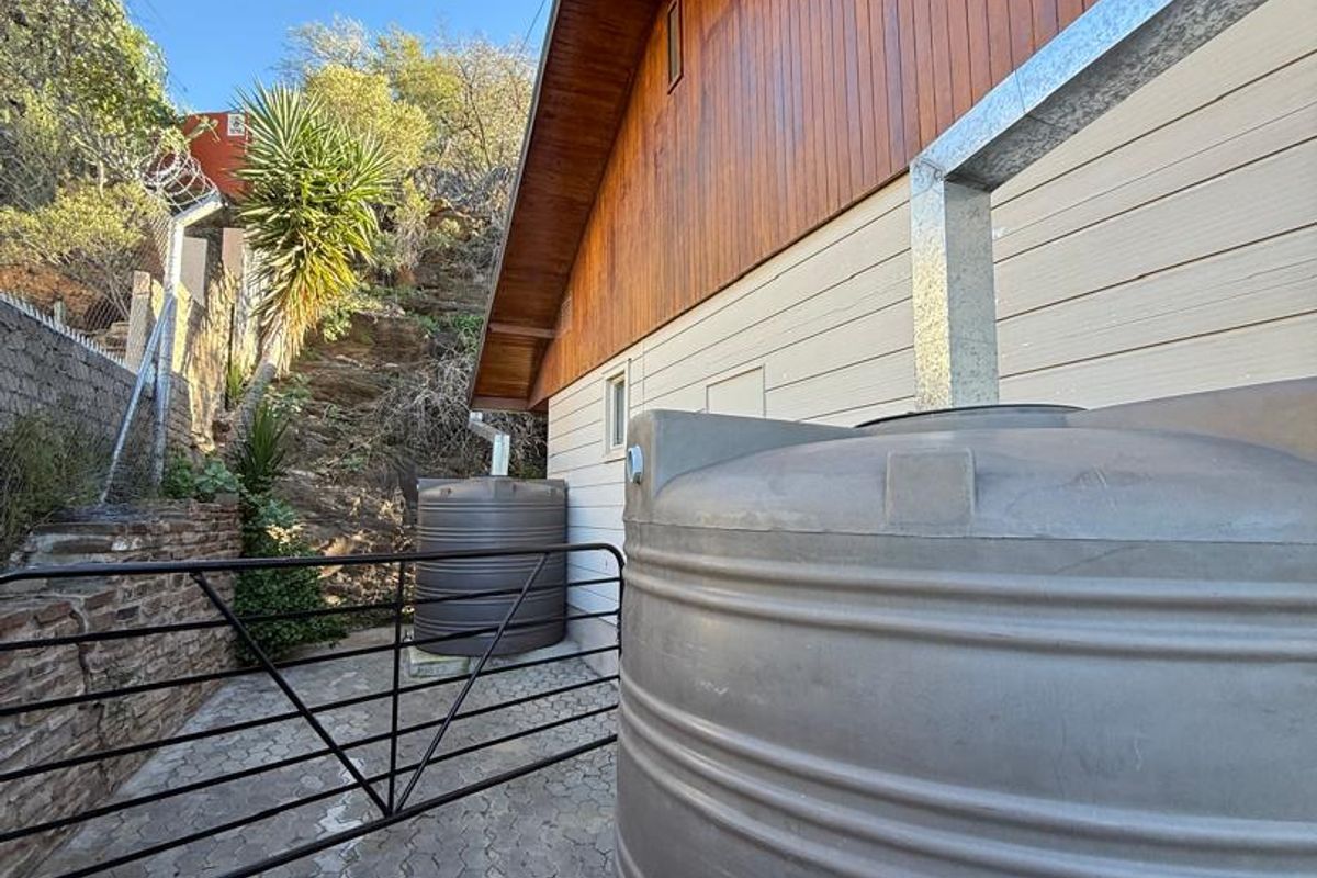 Rear exterior view featuring large water storage tanks, paved ground, and a metal security gate against a sloped hillside.