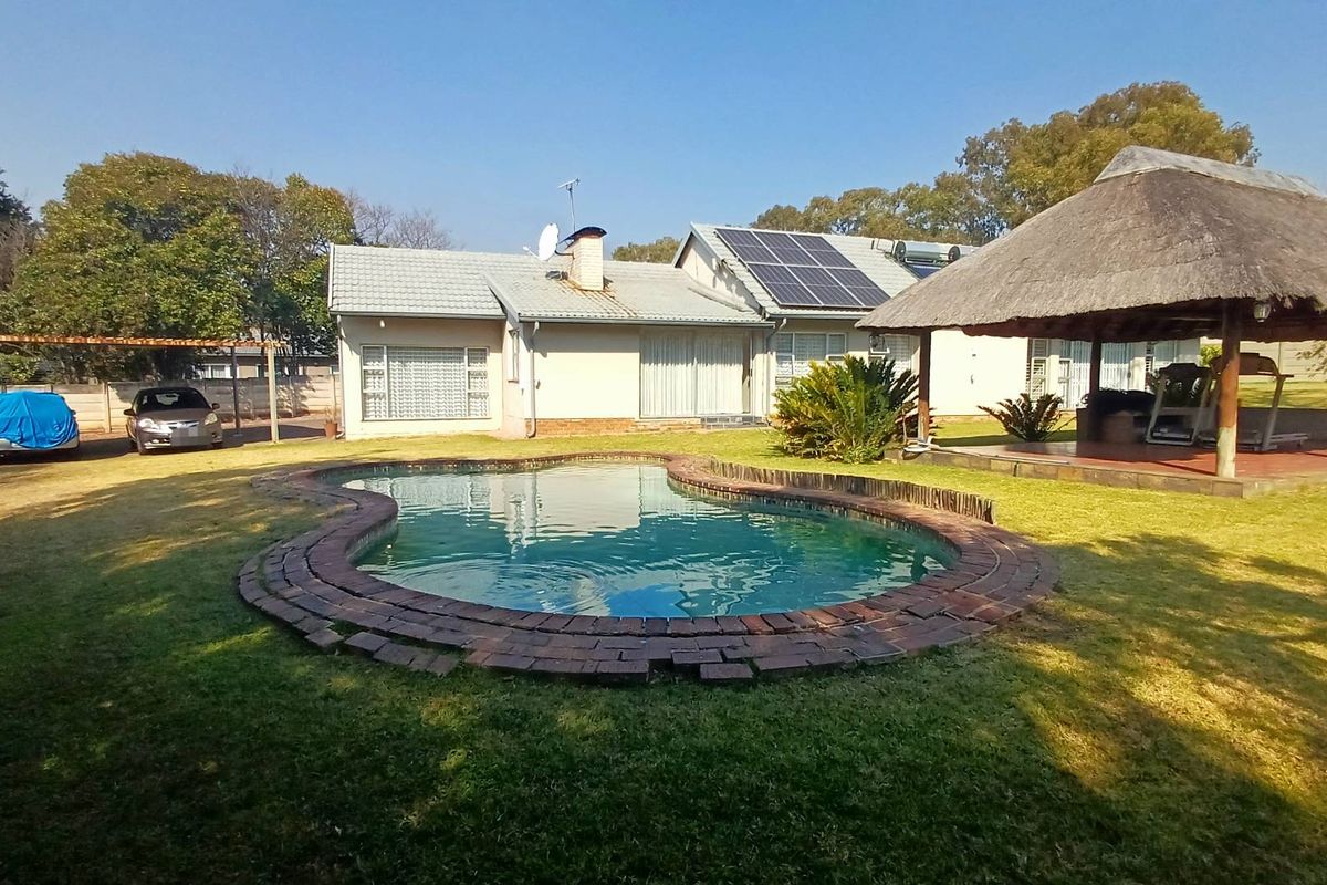 Rear garden view featuring a swimming pool with brick coping, a thatched lapa, solar panels on the roof, and a detached carport.
