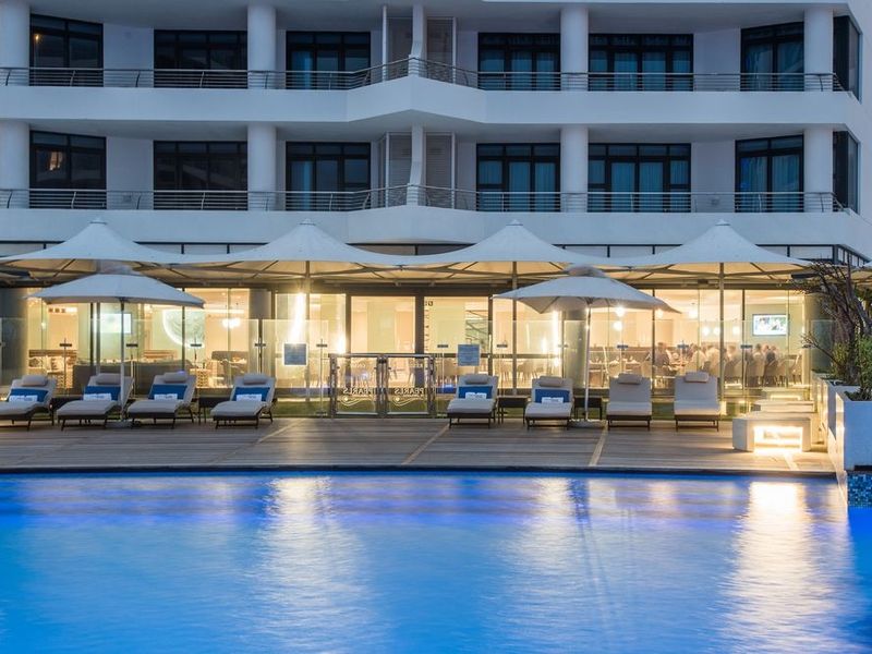 A well-maintained pool area featuring timber decking, glass balustrades, and modern tensile shade structures, set against a contemporary apartment building facade.