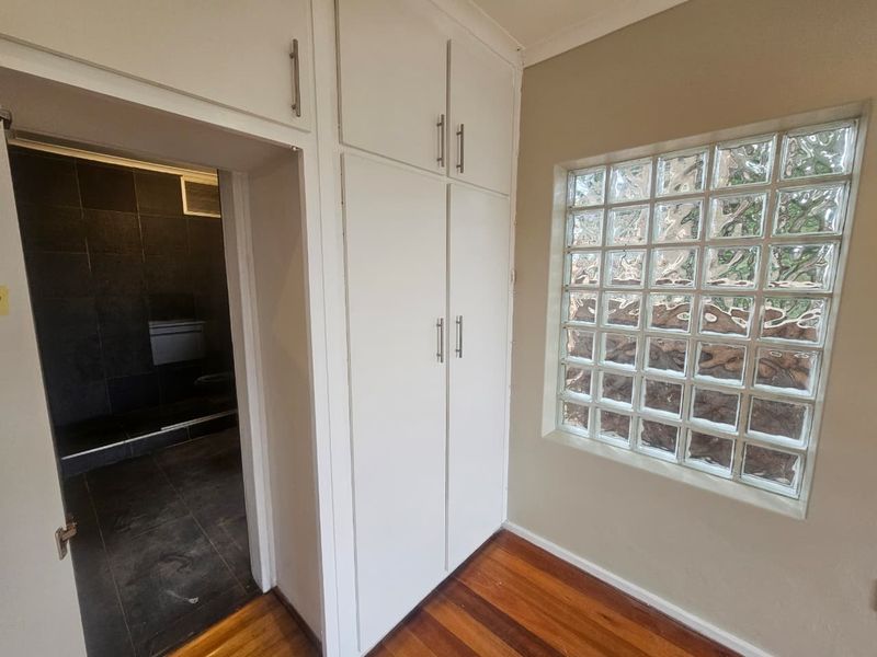 A bright hallway featuring built-in storage cupboards, hardwood flooring, and a decorative glass block window, providing access to an adjacent tiled bathroom.