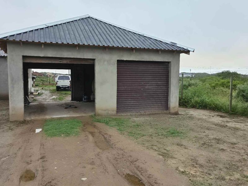 A detached garage structure featuring a corrugated metal roof, one roller shutter door, and an open bay, set on an unpaved plot.