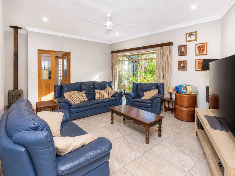 A comfortable living room featuring tiled flooring, a wood-burning fireplace, a ceiling fan, and large windows providing natural light and garden views.